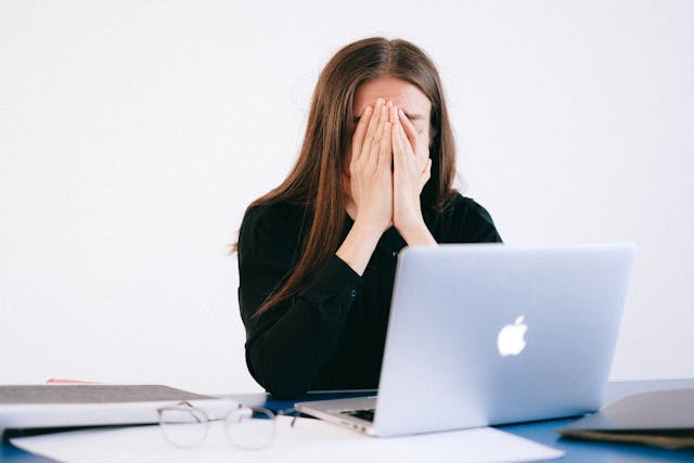 Frustrated woman sitting at a desk with her hands covering her face in front of a laptop, showing signs of burnout or work-related stress.