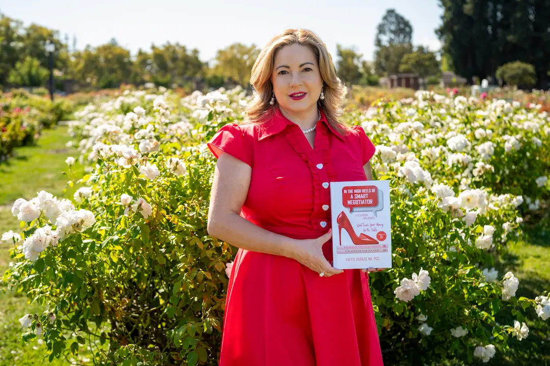 Yvette Durazo in a professional red dress holding her book in a white rose garden